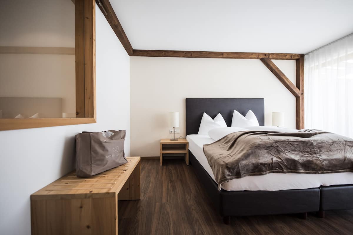 Un dormitorio con cabecera de madera, cama con almohadas blancas, mesita de noche con lámpara y banco de madera.