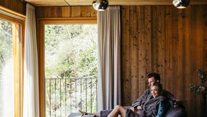 A modern living room with a wooden ceiling, a large window with curtains, and a sofa with cushions. - Hotel Bad Fallenbach (San Leonardo in Passiria)
