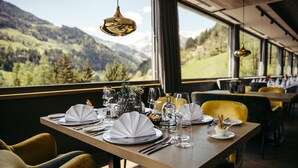 A dining table set with plates, cutlery, and glasses, with a mountain view through large windows. - Hotel Bad Fallenbach (San Leonardo in Passiria)