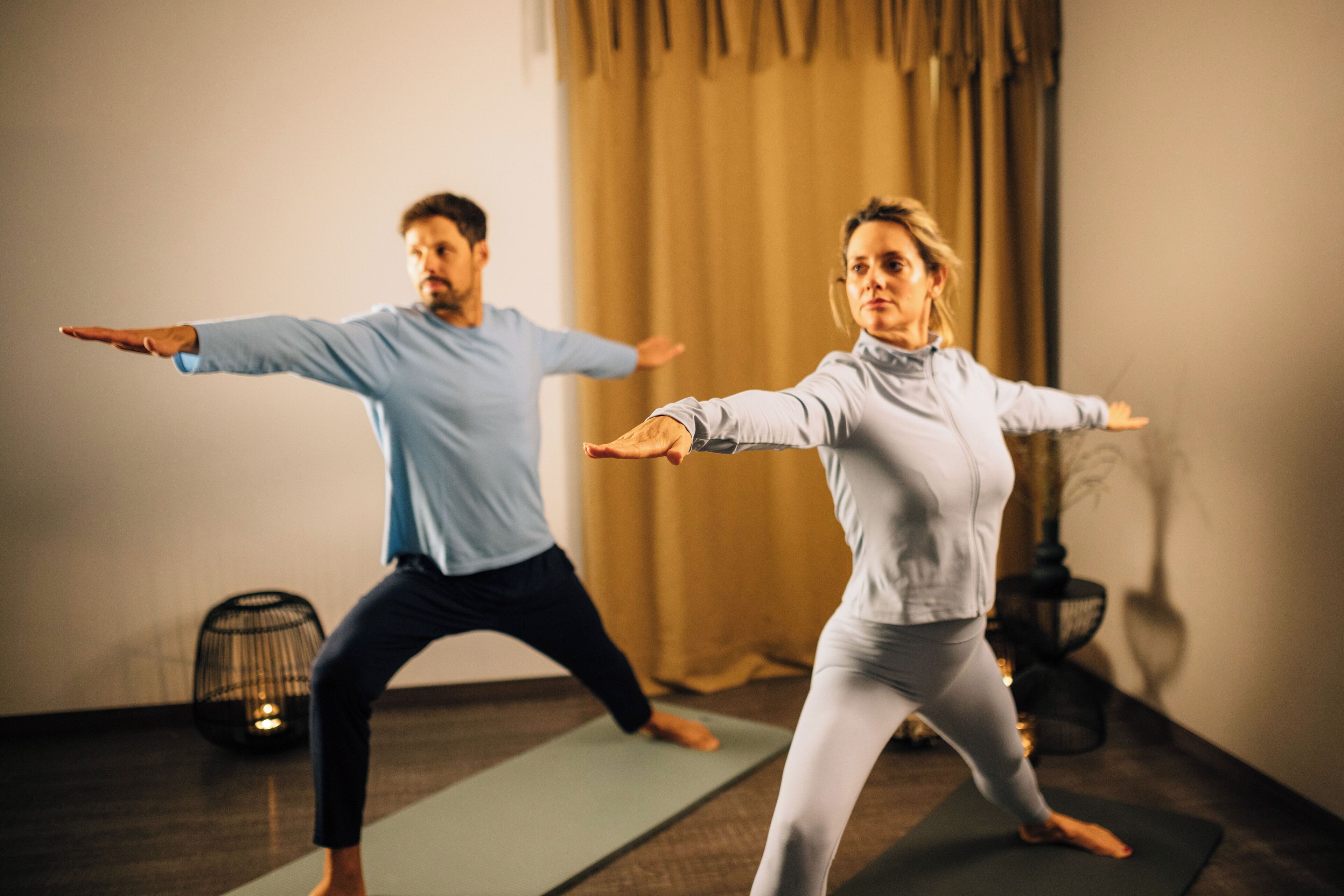 Two people practicing yoga in a room with a large window and curtains.
