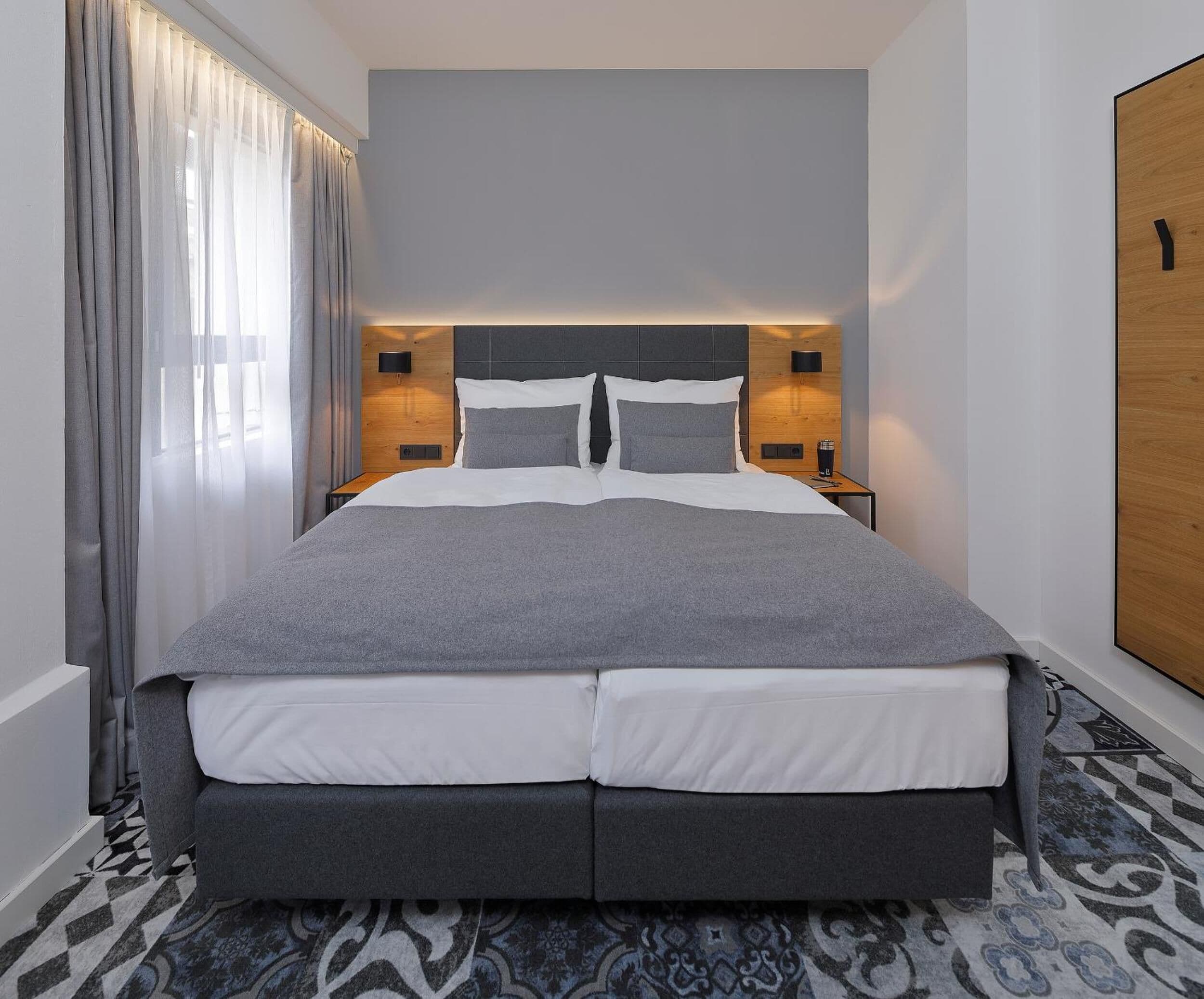 Ein modernes Hotelzimmer mit einem großen Bett, grau gestreuter Bettwäsche, einem gemusterten Teppich und einem Holzschrank.