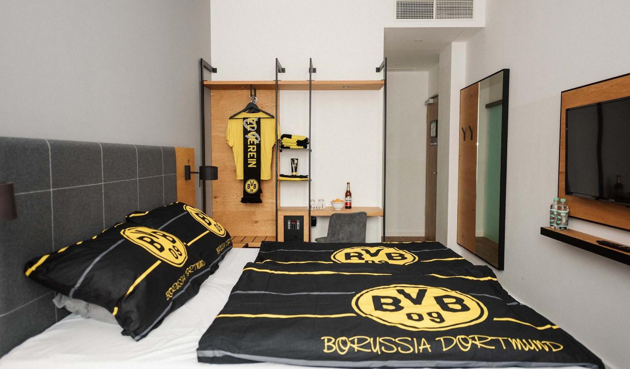 A hotel room with two beds, a wooden headboard, a small desk with a chair, a mirror, and a shelf with toiletries.