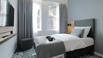 A modern hotel room with a bed, a TV, a window with curtains, and a 'Feel at Home' sign on the bed.