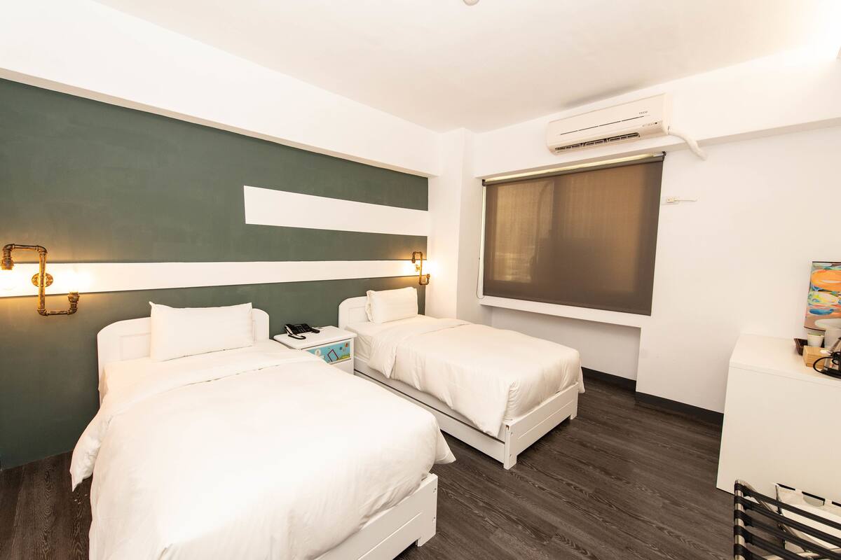 Habitación de hotel con dos camas, un escritorio, una silla, una ventana y un equipo de aire acondicionado.