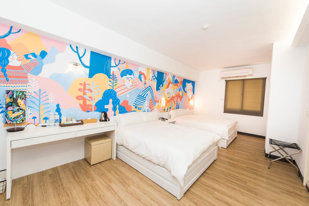 Habitación de hotel moderna con un mural grande y colorido, dos camas, un escritorio y un pequeño rincón de descanso.