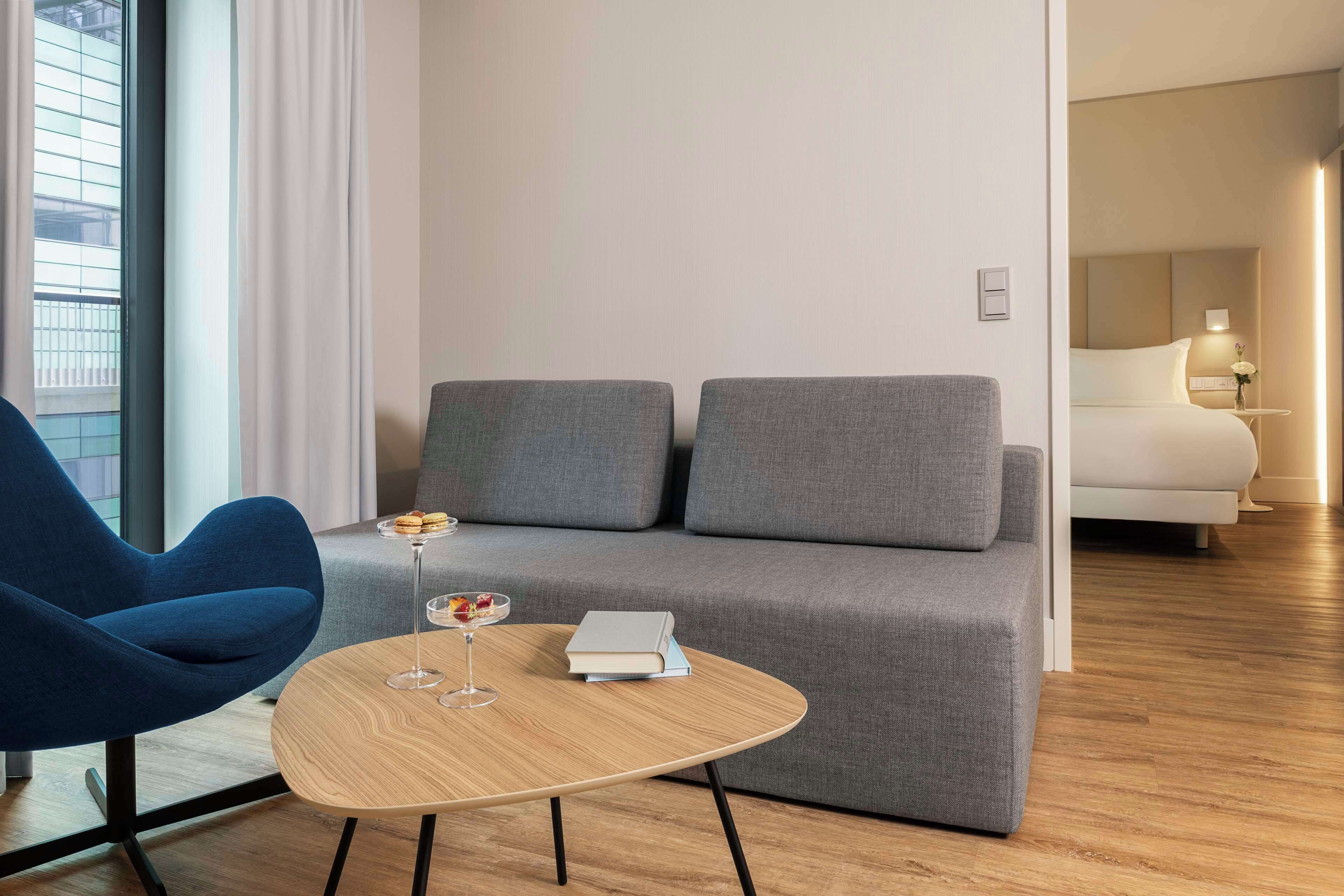 Una habitación de hotel moderna con un sofá gris, un sillón azul, una mesa de centro de madera con un libro y un vaso de bebida, y una cama con una mesita de noche.