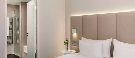 A modern hotel room with a large bed, a bedside table with a vase, and a view into the bathroom.