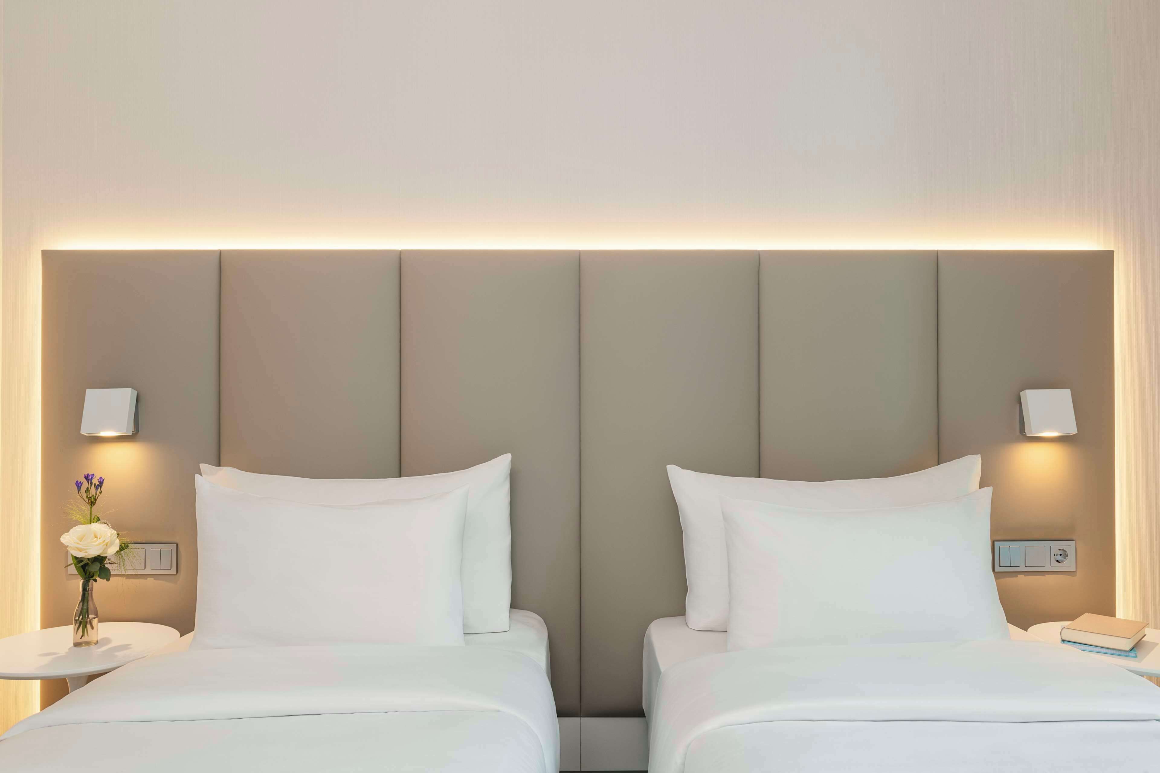 Una habitación de hotel con dos camas, una mesita de noche con lámpara, y una lámpara fijada a la pared.