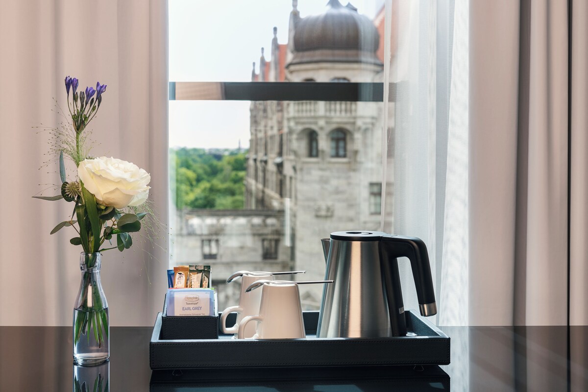 A hotel room with a view of a historic building, a vase with flowers, a coffee maker, and a tray with a mug and a box of toiletries.