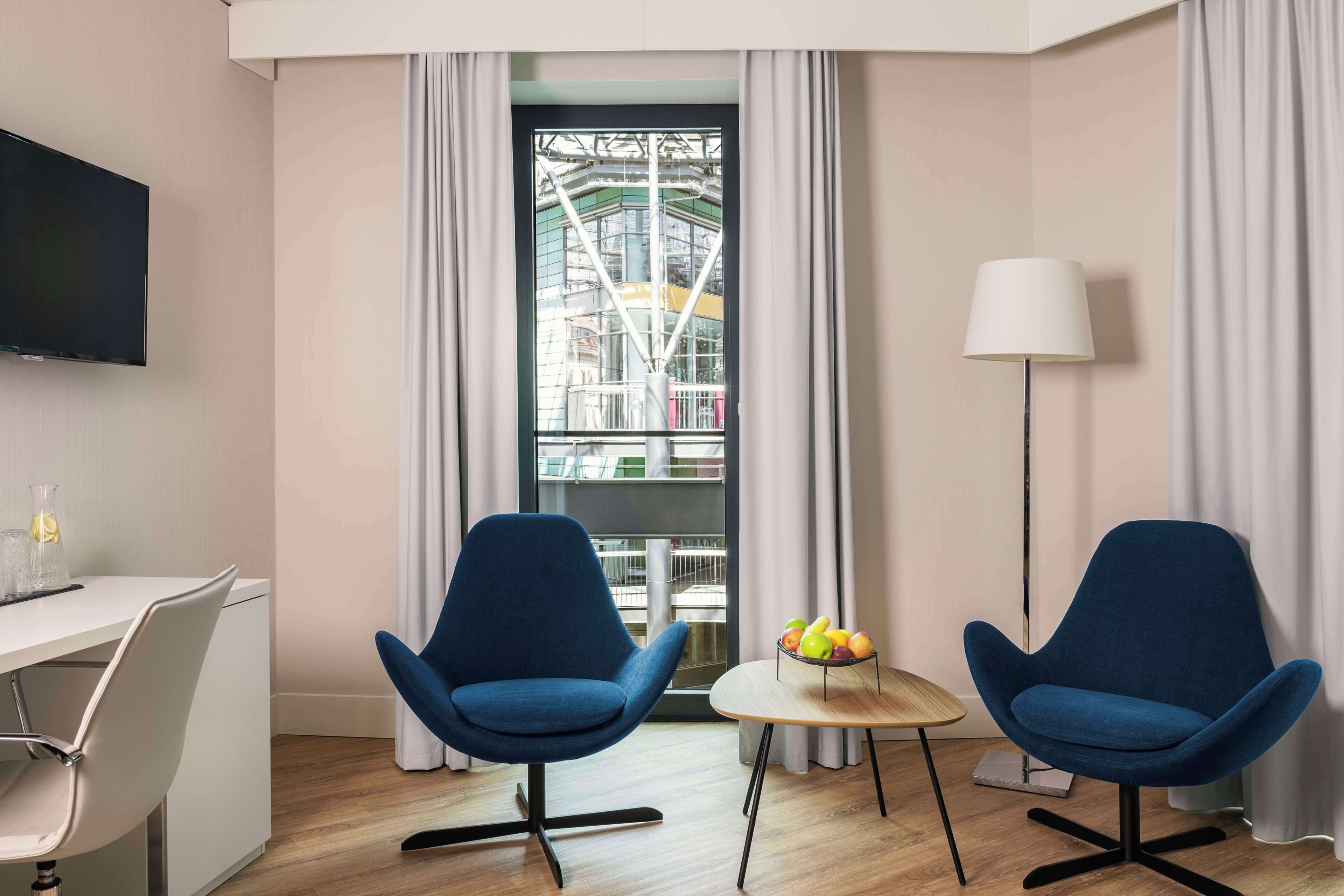 A modern hotel room with two blue armchairs, a small round table with fruit, a desk with a lamp, and a large window with a view of a building.