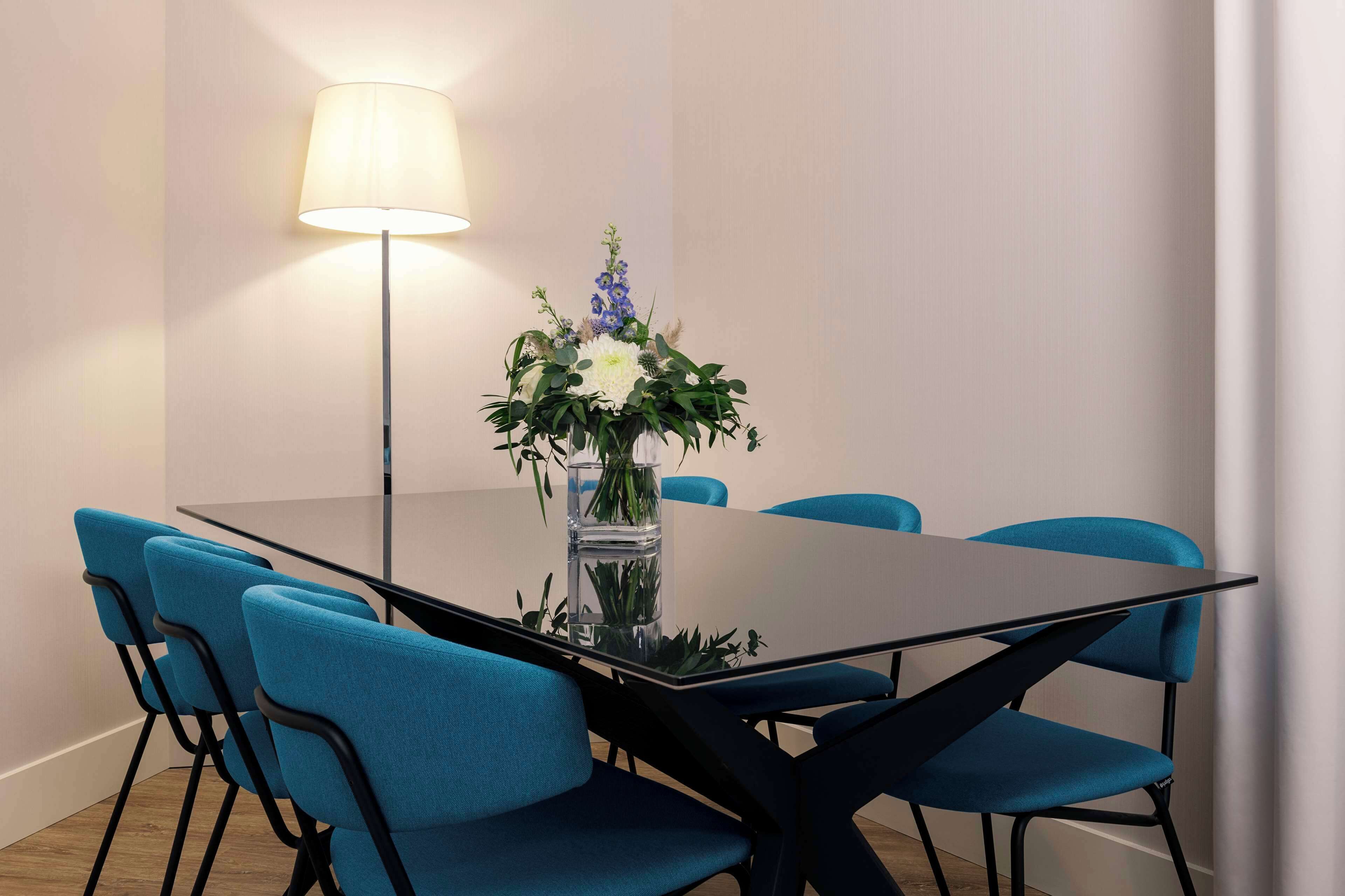A conference table with a vase of flowers, blue chairs, and a floor lamp.