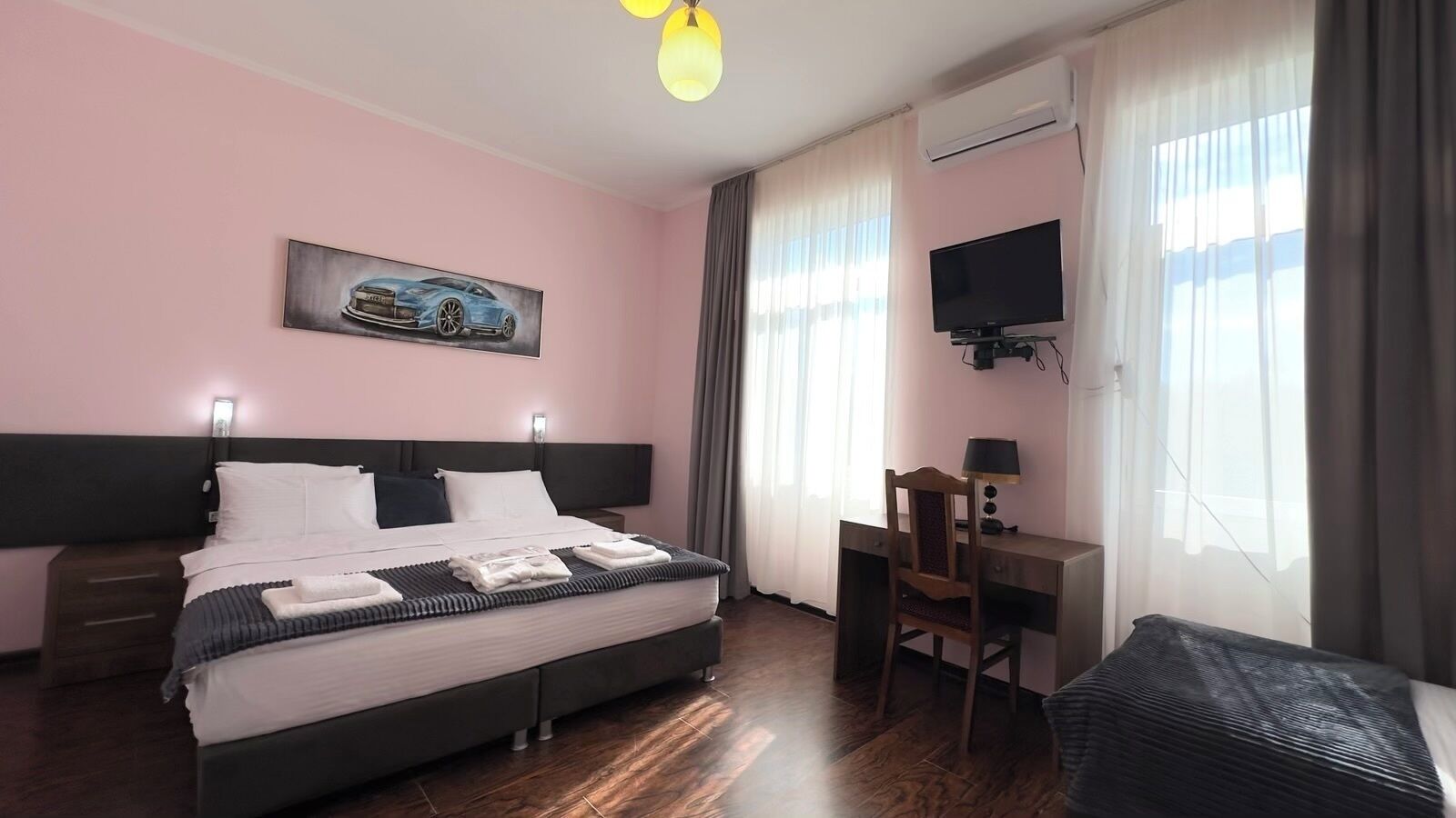 A hotel room with a bed, a desk, a chair, a TV, and a painting on the wall.