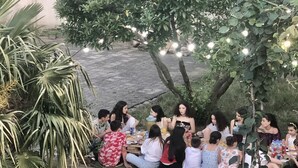Un grupo de personas sentadas en un césped bajo un árbol con luces colgantes, rodeado de follaje.
