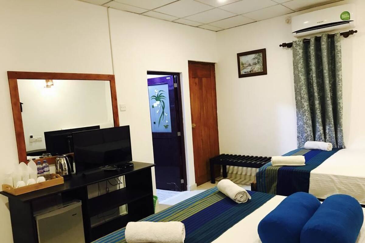 A hotel room with a TV, a desk with a computer, a bed with blue pillows, and a window with curtains.