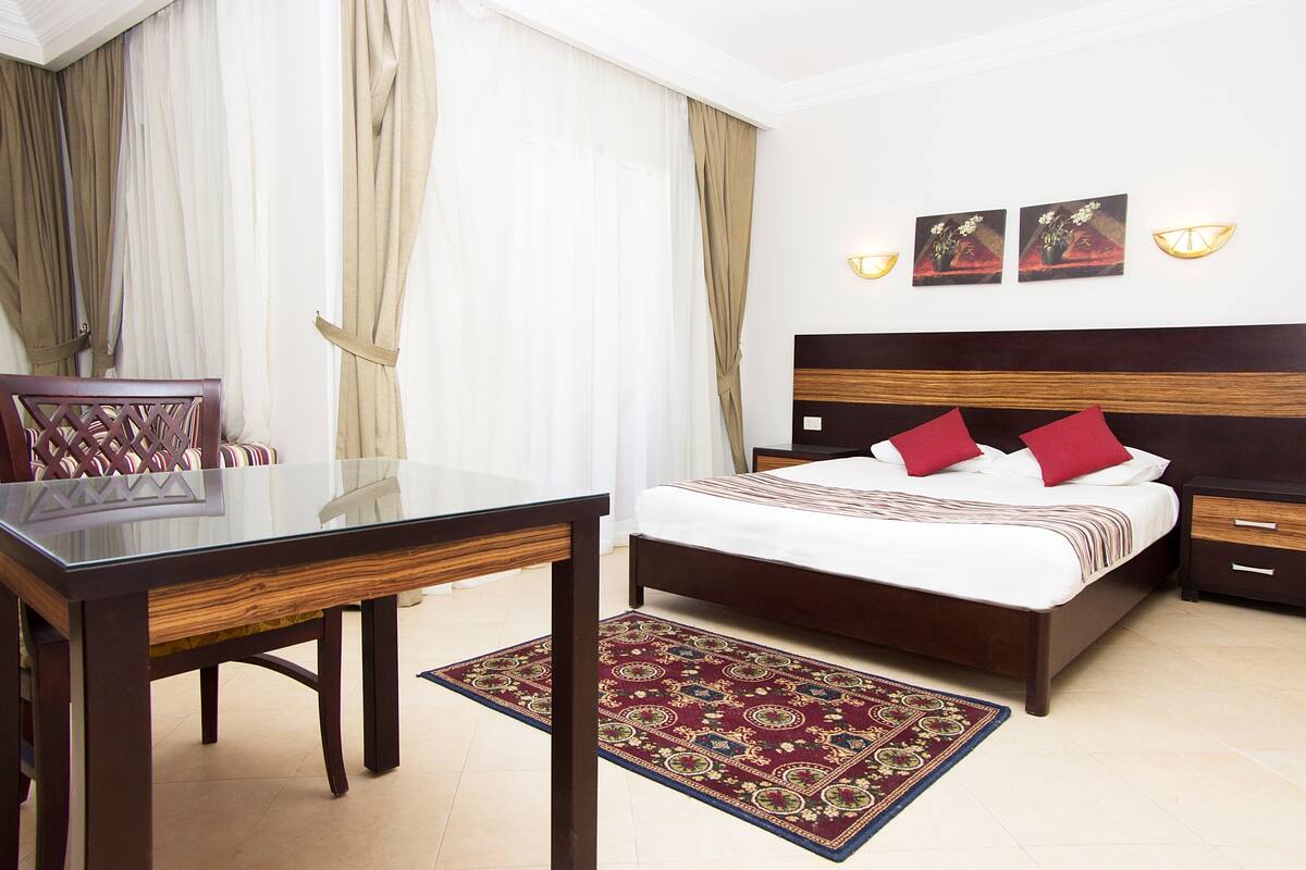 A hotel room with a wooden bed, a glass table, a red and gold patterned rug, and a nightstand with a lamp.