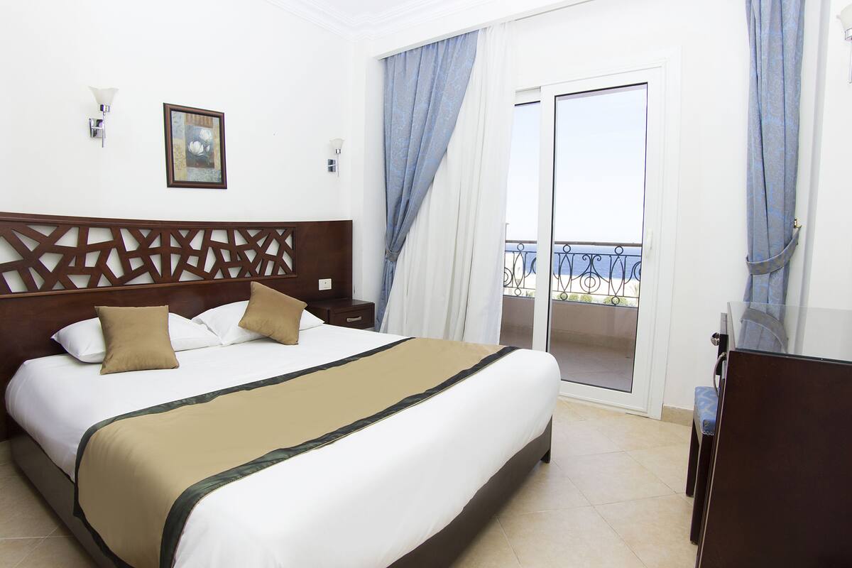 A hotel room with a large bed, a wooden headboard, a nightstand, a desk, and a balcony with a view.