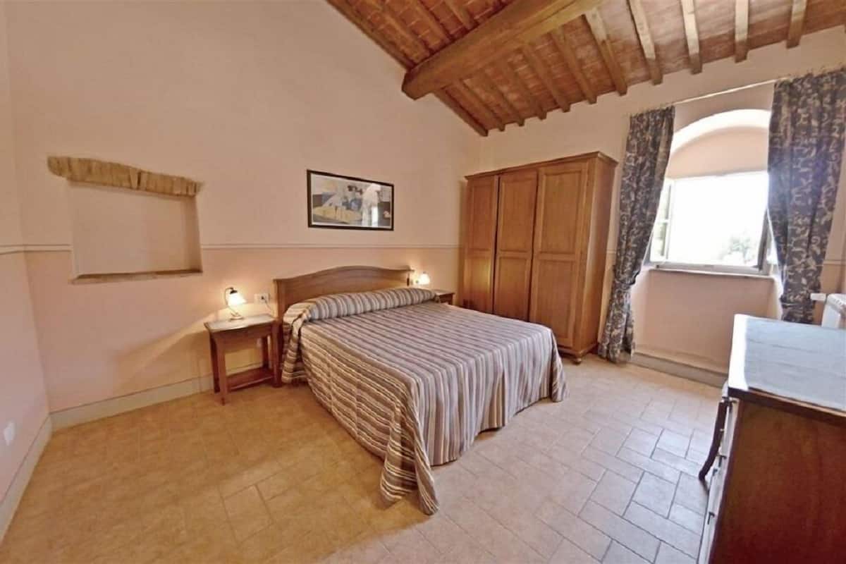 A bedroom with a bed, wooden wardrobe, bedside table, lamp, and a window with curtains.