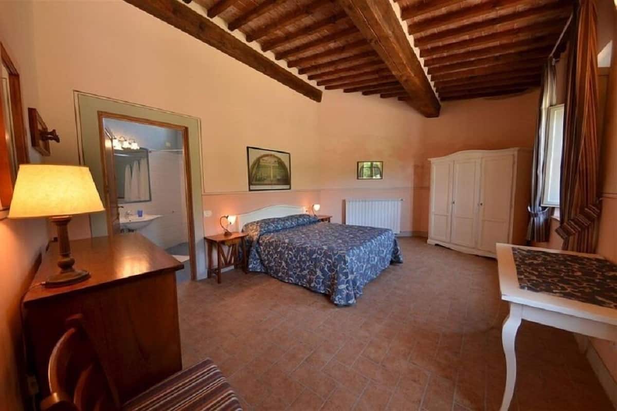 A bedroom with a bed, a nightstand, a lamp, a radiator, a wardrobe, and a window with curtains.