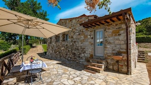 A stone building with a small porch, a table with chairs, and a table umbrella. - Agriturismo Concadoro (Castellina in Chianti)