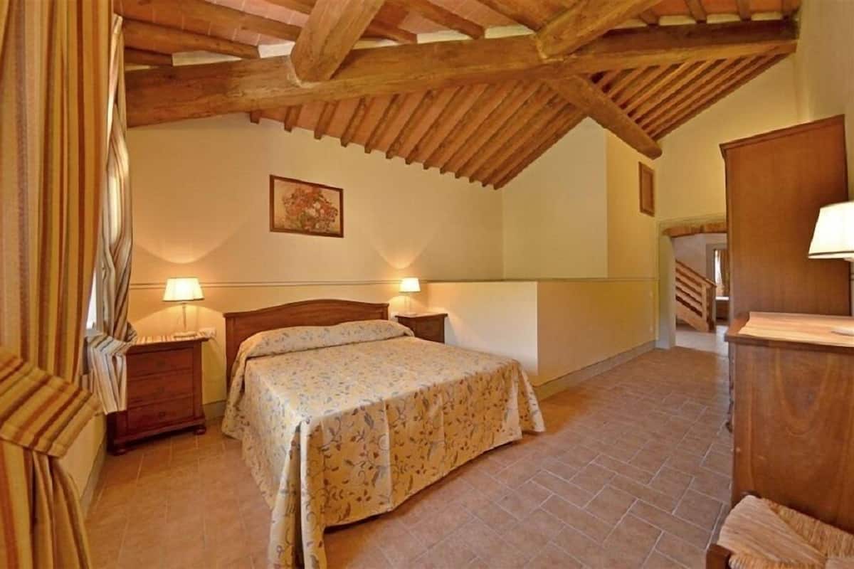 A bedroom with a wooden ceiling, a bed with a floral bedspread, two bedside lamps, a wooden nightstand, and a wooden wardrobe.