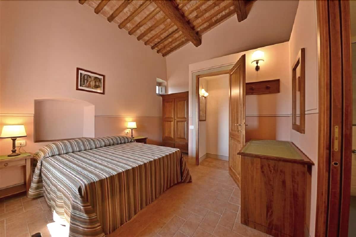 A bedroom with a bed, two bedside lamps, a wooden wardrobe, and a door leading to another room.