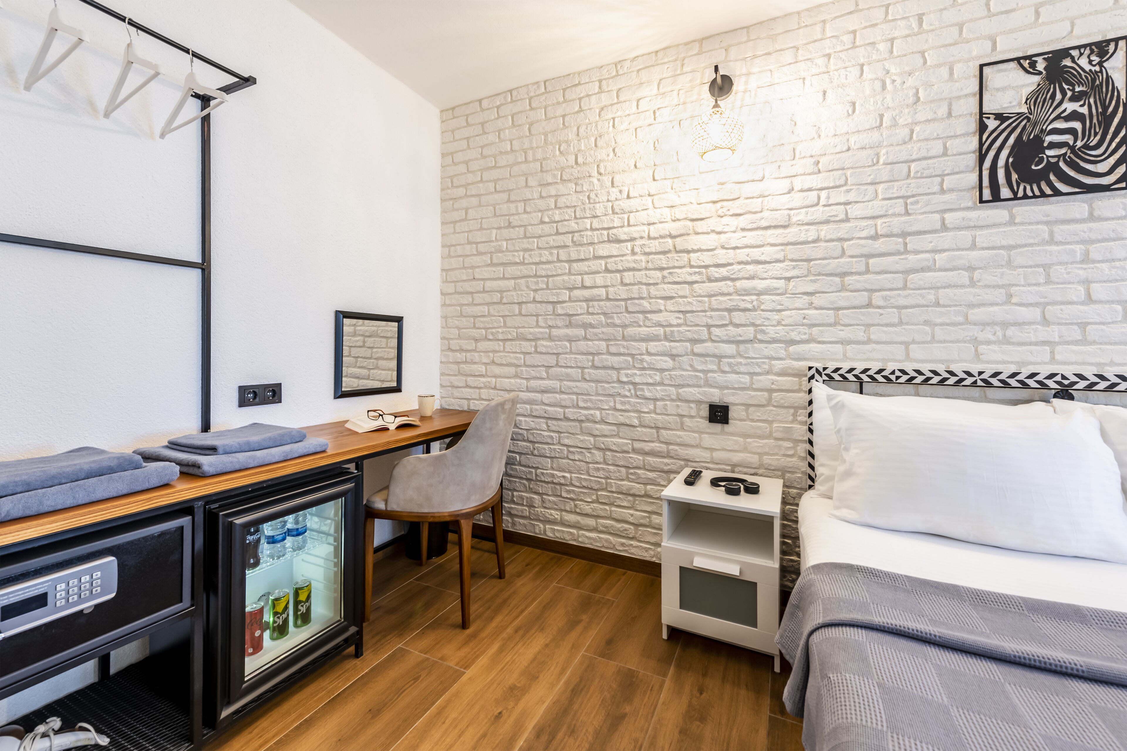 Une chambre d’hôtel compacte comprenant un lit, un bureau avec une chaise, un petit réfrigérateur, une table de chevet et un mur en briques blanches.