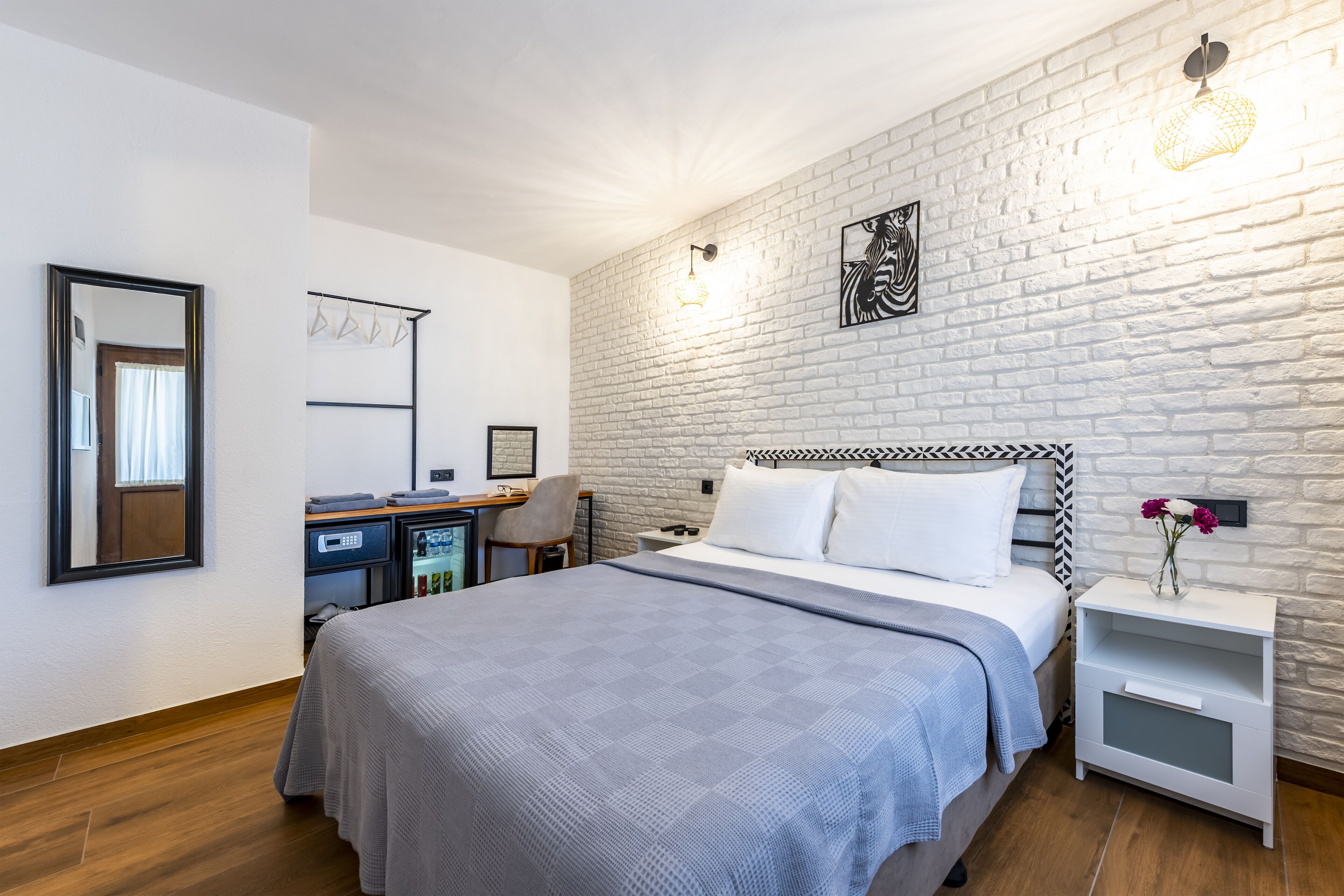 A modern hotel room with a large bed, a desk with a chair, a wardrobe, and a white brick wall.