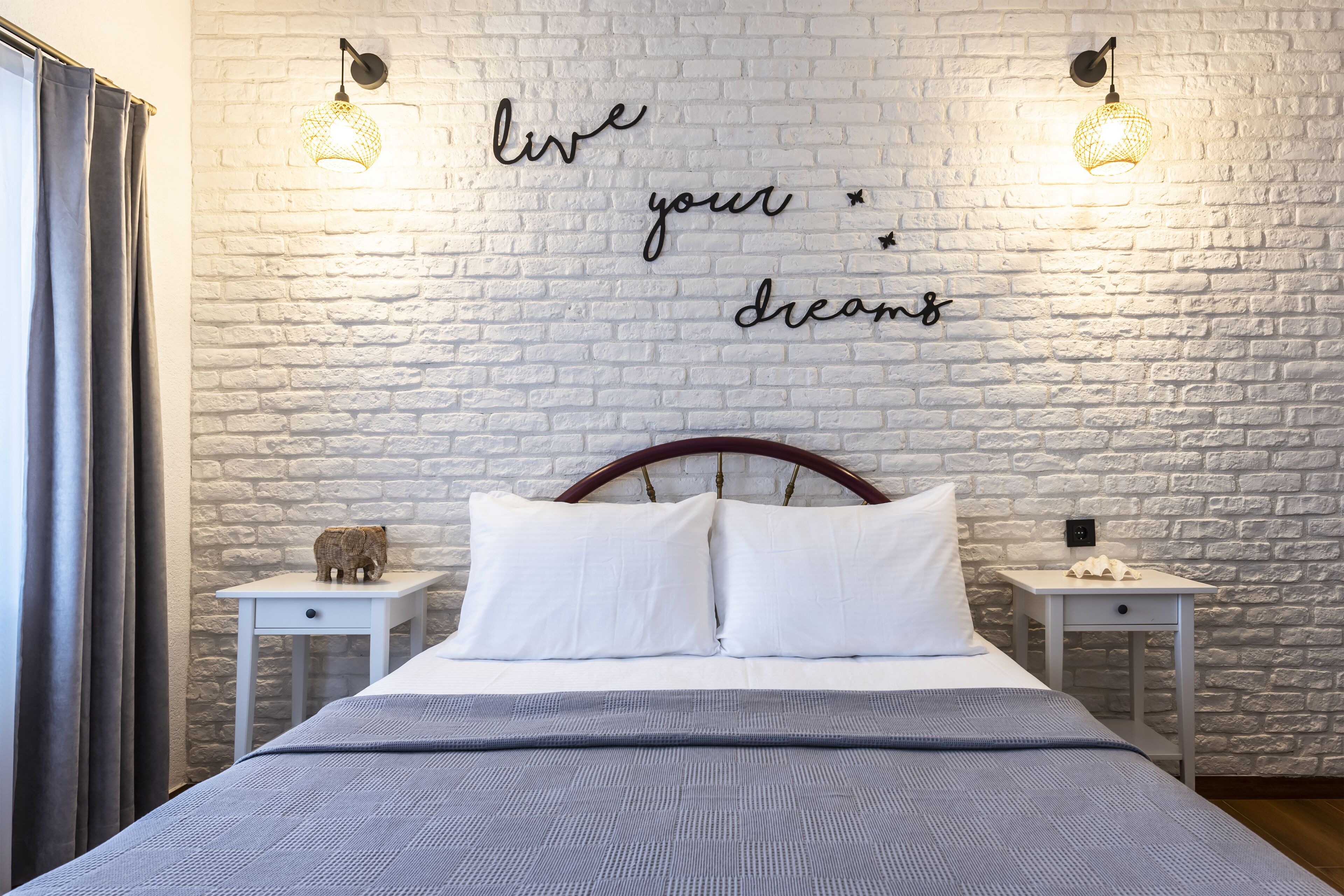 Una cama bien tendida con sábanas blancas, dos mesitas de noche con adornos, y una pared de ladrillo blanco con texto inspirador.