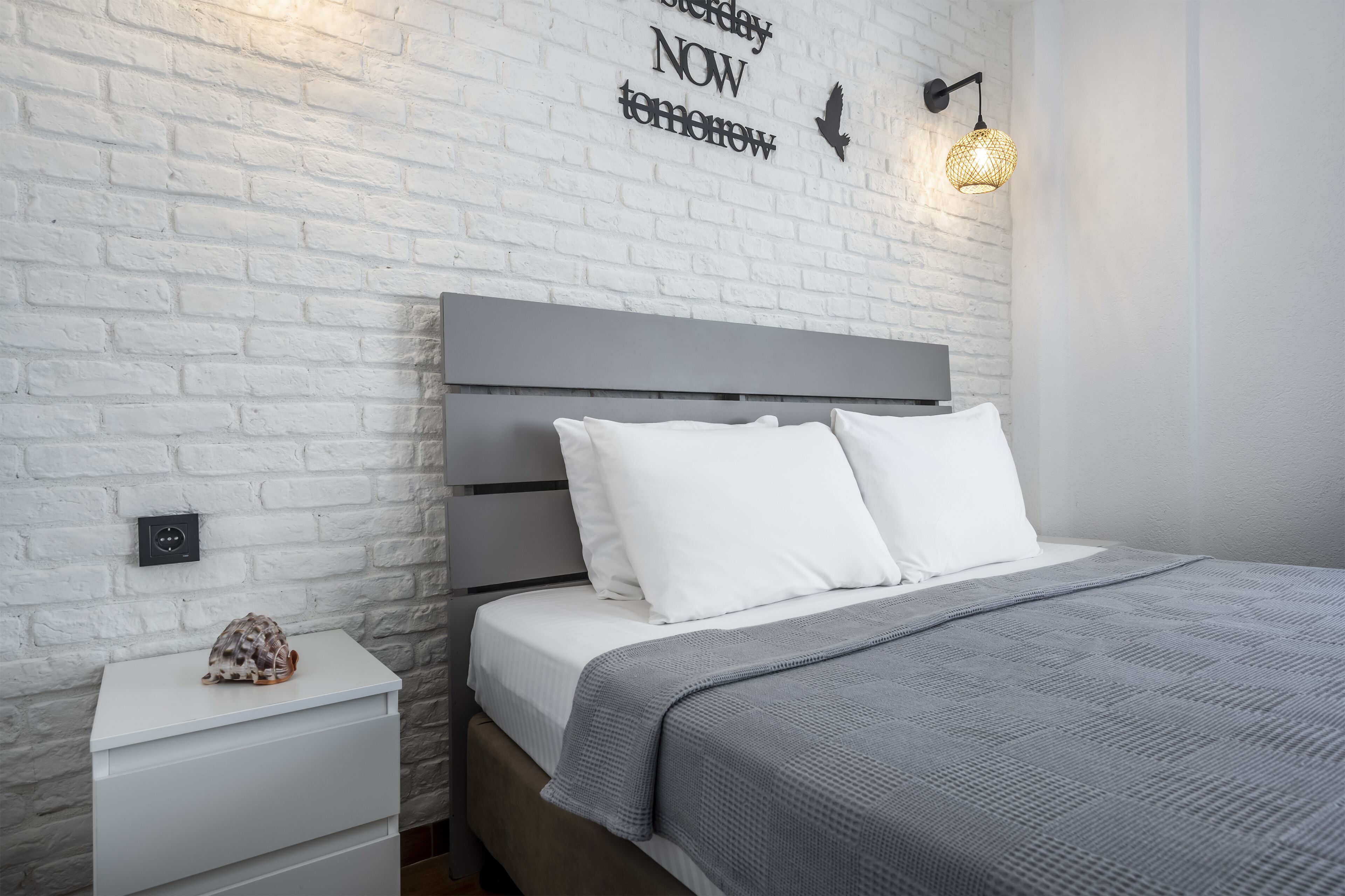 Un dormitorio moderno con pared de ladrillo blanco, cabecero gris, ropa de cama blanca y manta gris.