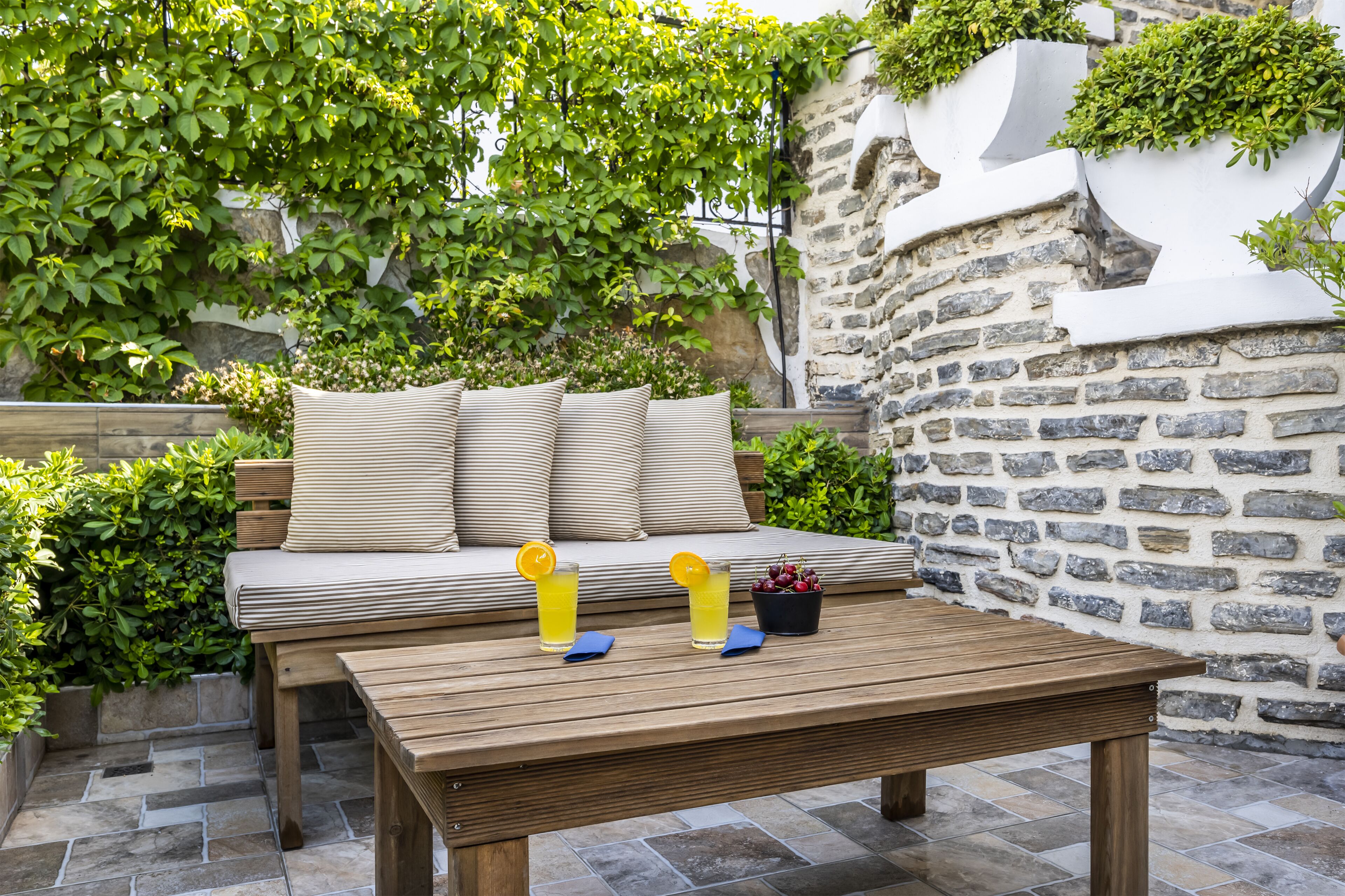 Una mesa de madera con bebidas y un cuenco de fruta, un banco acolchado con cojines a rayas, y una pared de piedra con plantas.