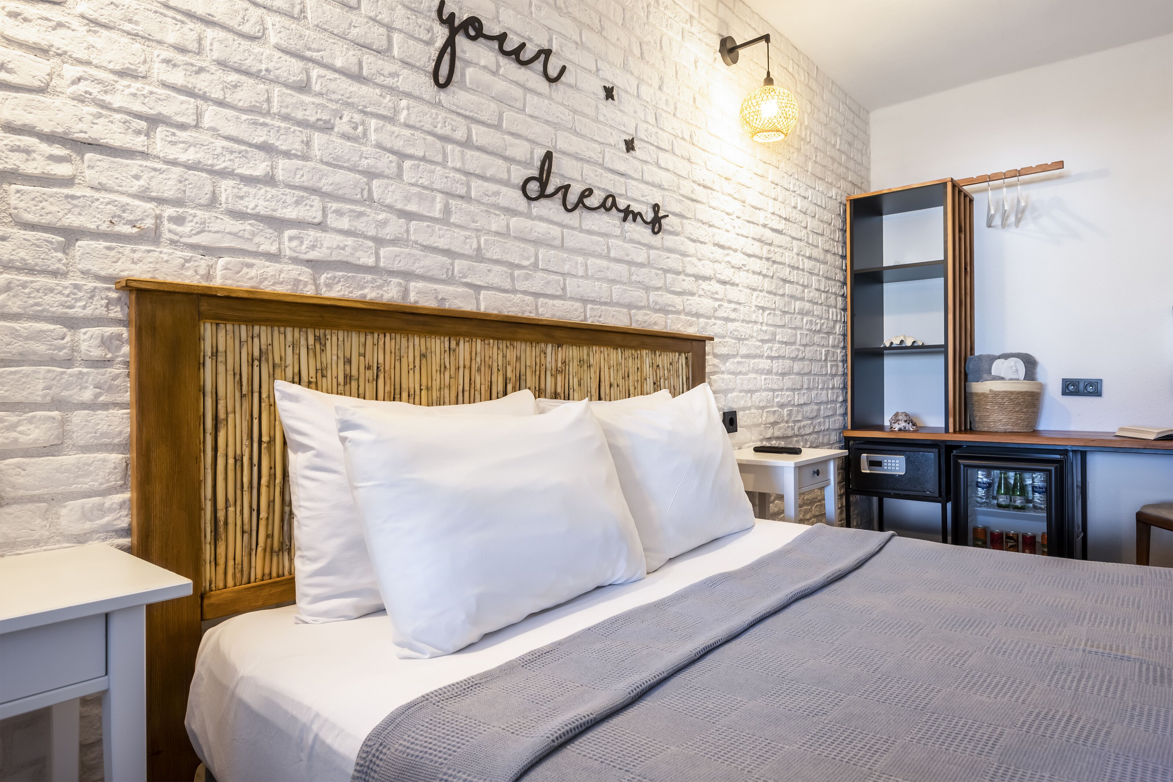 A neatly made bed with white linens and a grey blanket, a wooden headboard, a wall with a white brick pattern, and a shelf with various items.