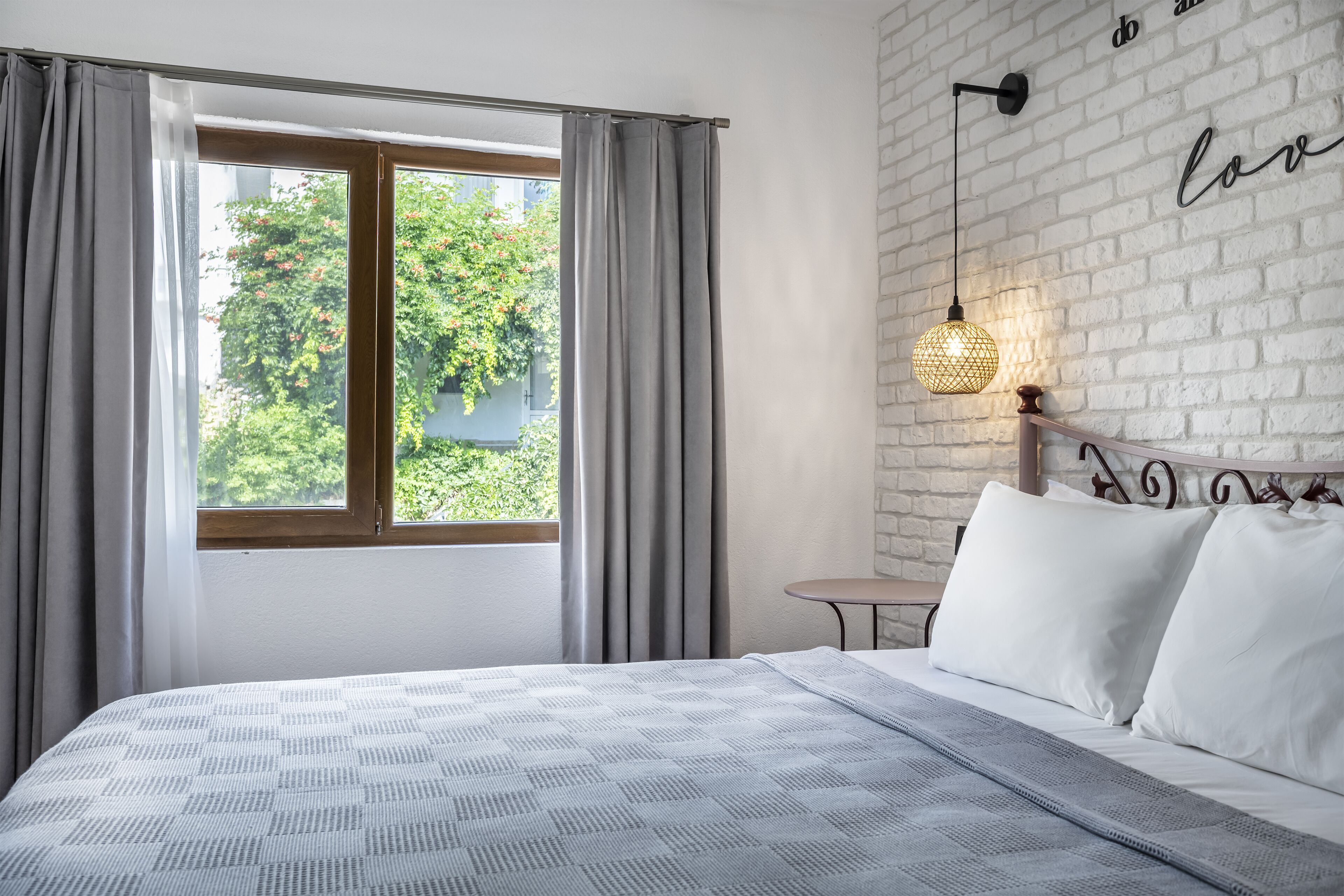 A bedroom with a large bed, a window with curtains, a small round table, and a wall with a brick pattern and a 'do' and 'love' sign.