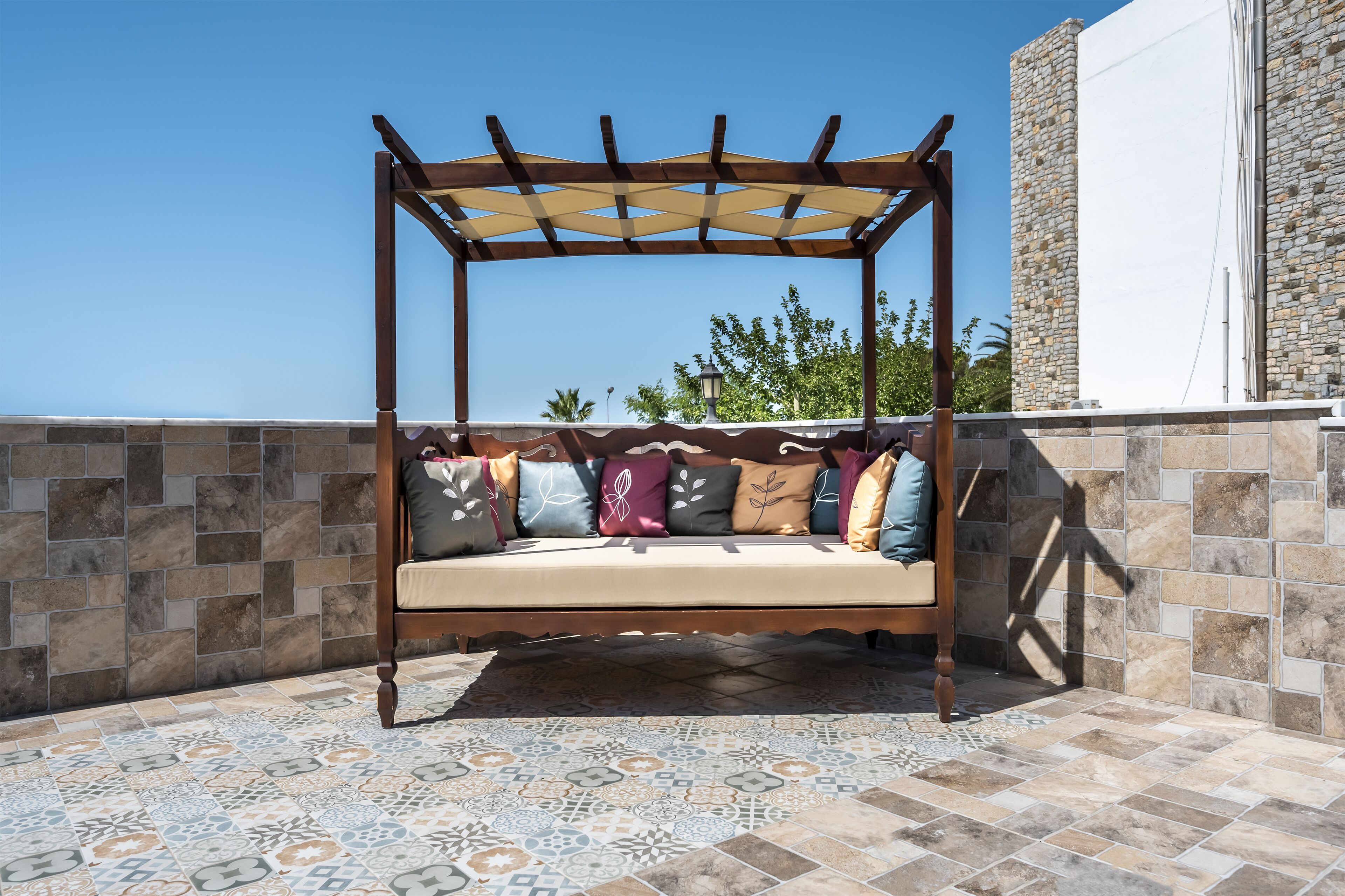 Un espacio de estar al aire libre con madera, que incluye un toldo, un banco acolchado y cojines con motivos, además de un suelo de baldosas.