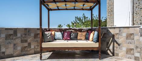Un espacio de estar al aire libre con madera, que incluye un toldo, un banco acolchado y cojines con motivos, además de un suelo de baldosas.