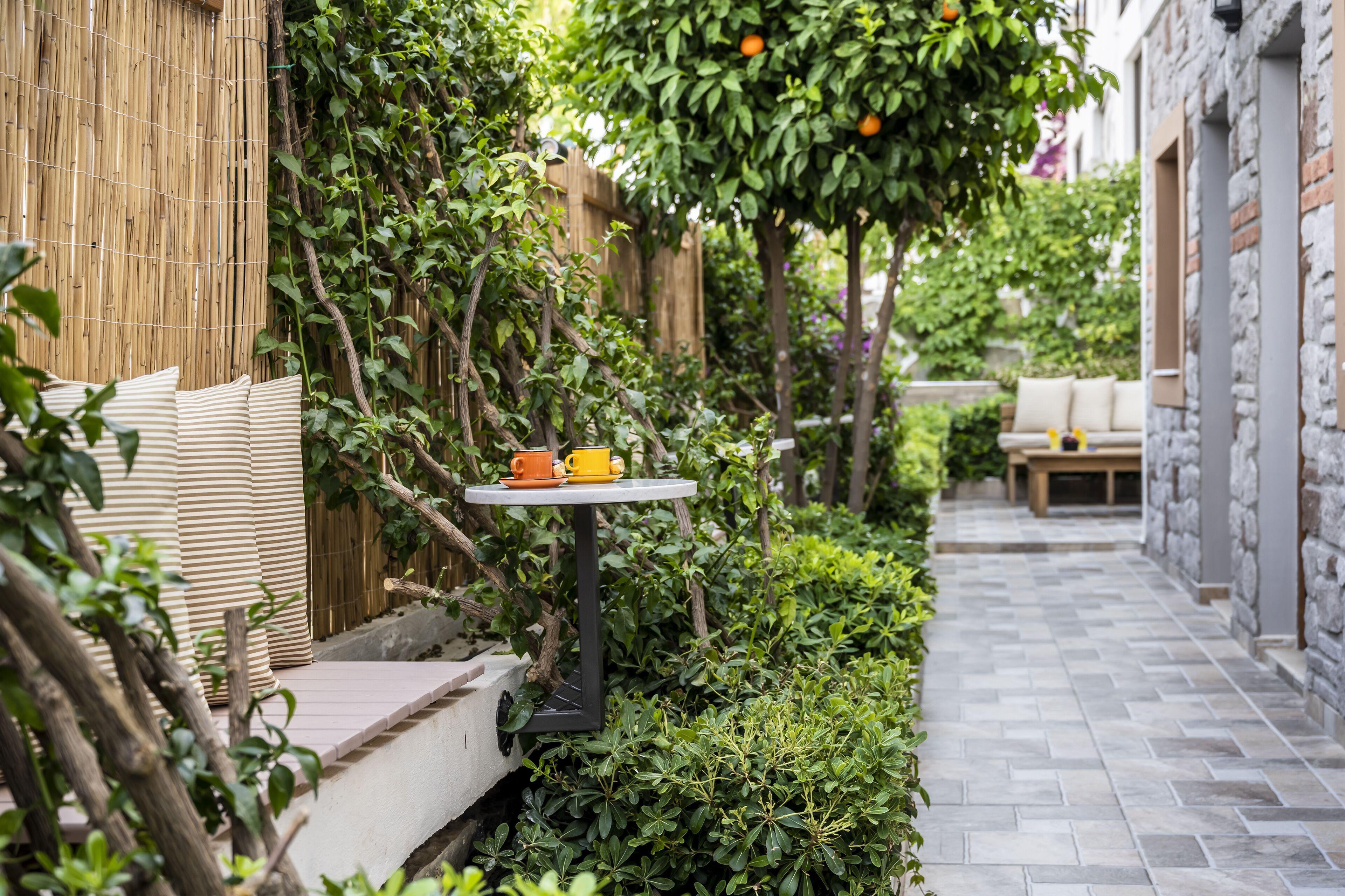A narrow outdoor pathway with a small table and chairs, surrounded by lush greenery and a bamboo fence.
