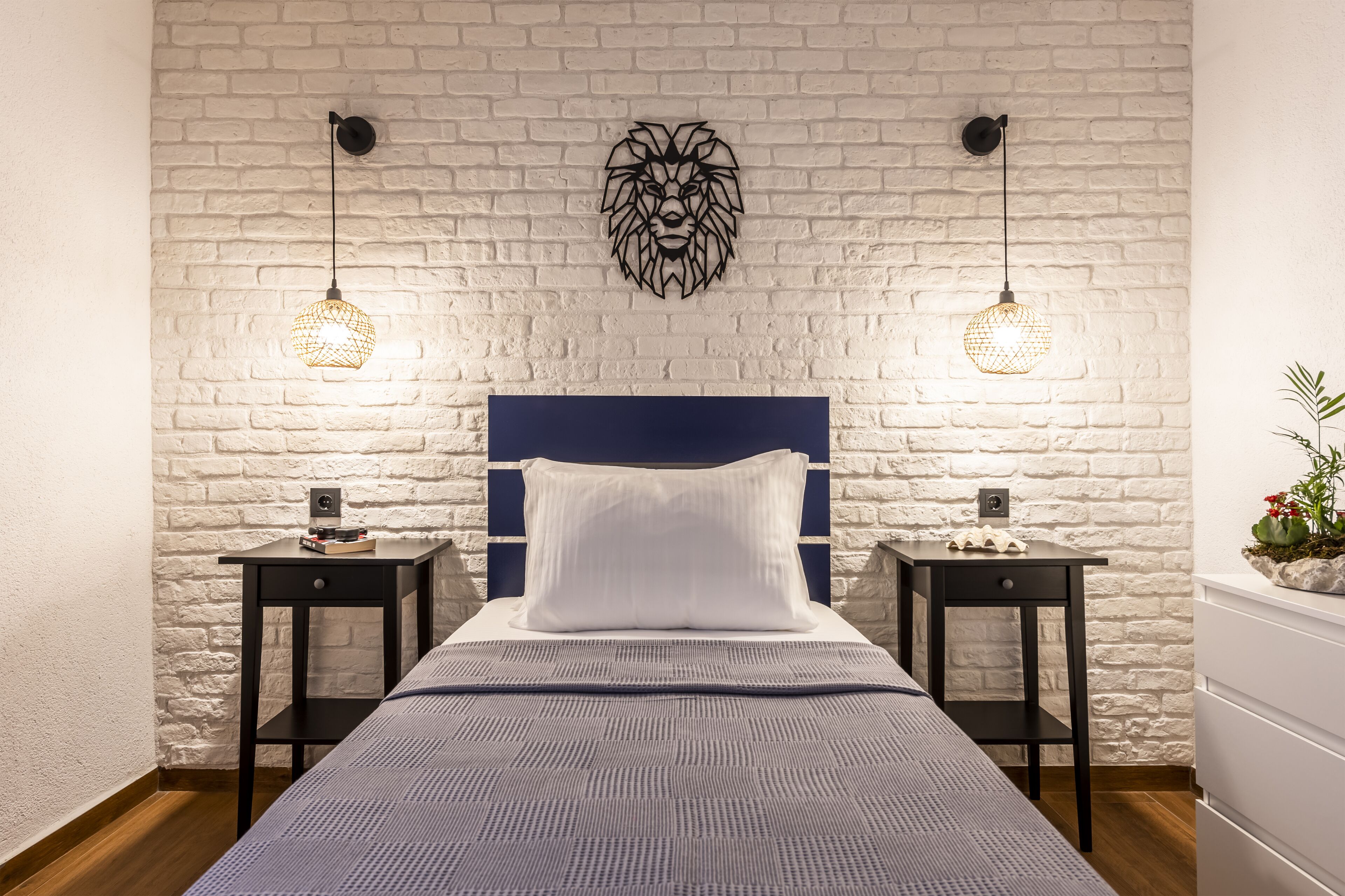 A neatly arranged bedroom with a brick-patterned wall, a blue headboard, two bedside tables with lamps, and a decorative wall piece above the bed.