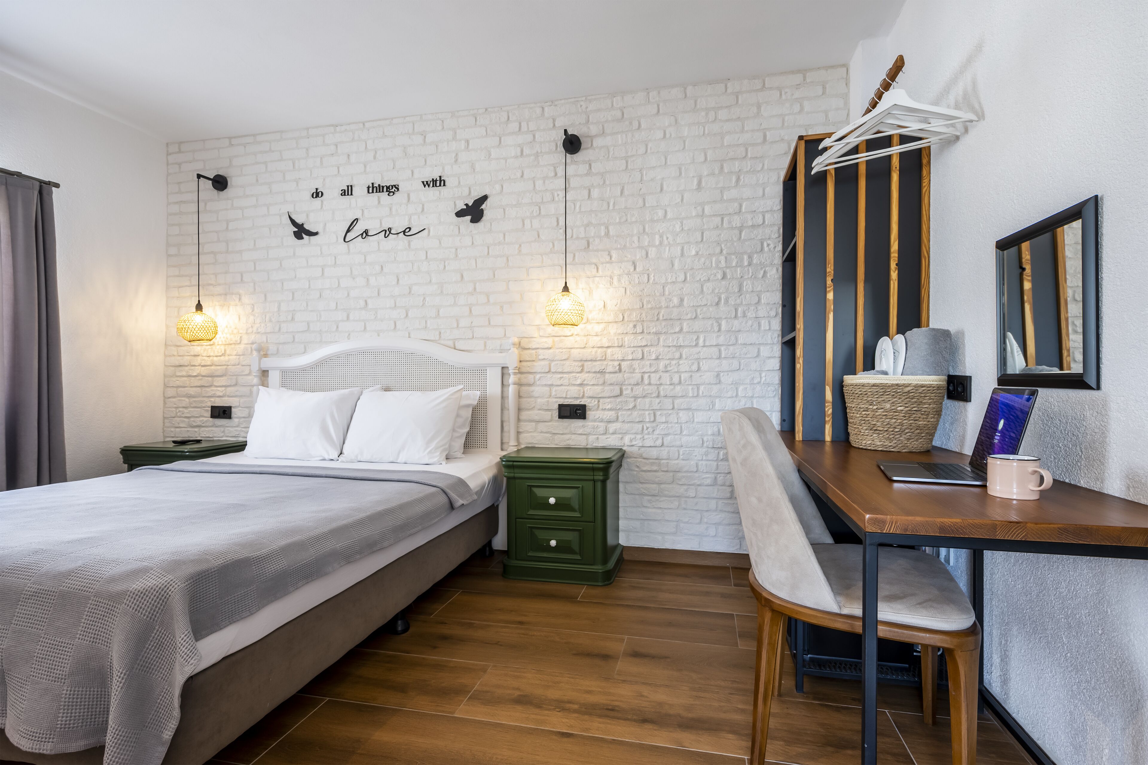 Un dormitorio con pared de ladrillo blanco, cama con ropa de cama blanca, mesita de noche verde, escritorio de madera con una computadora portátil y una taza, y un espejo en la pared.