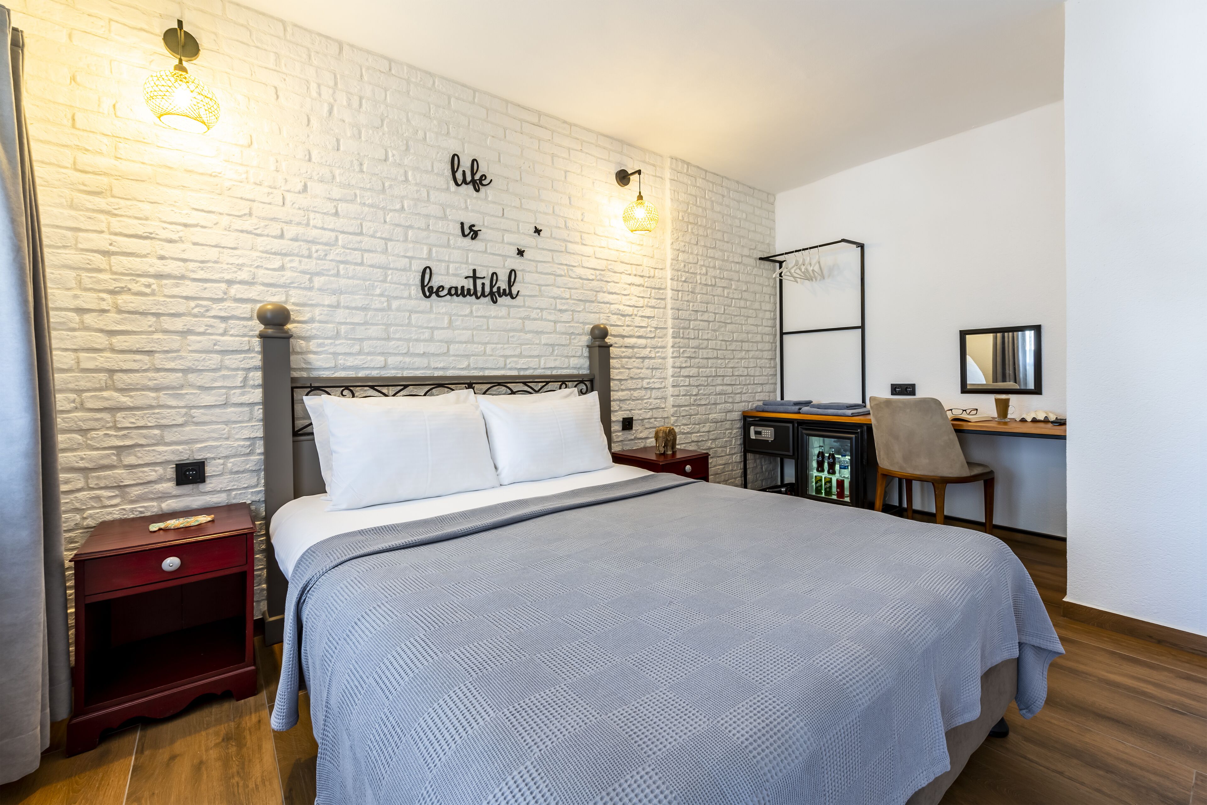 A bedroom with a bed, a nightstand, a desk, and a chair. There is a white brick wall with a sign that says "life is beautiful".