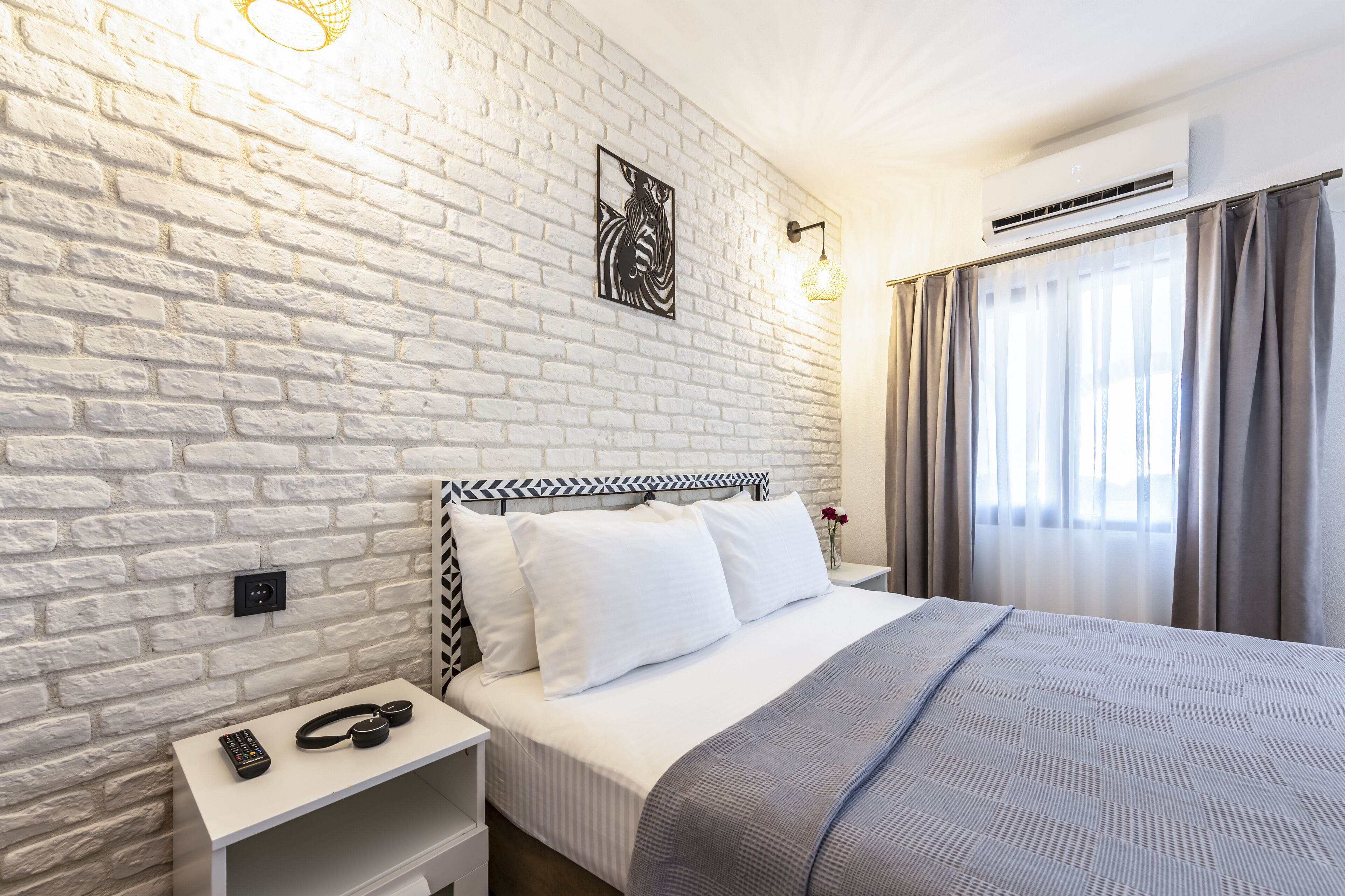 Ein Hotelzimmer mit weißer Ziegelwand, einem Bett mit weißer Bettwäsche, einem Nachttisch mit Fernbedienung und einer Klimaanlage.