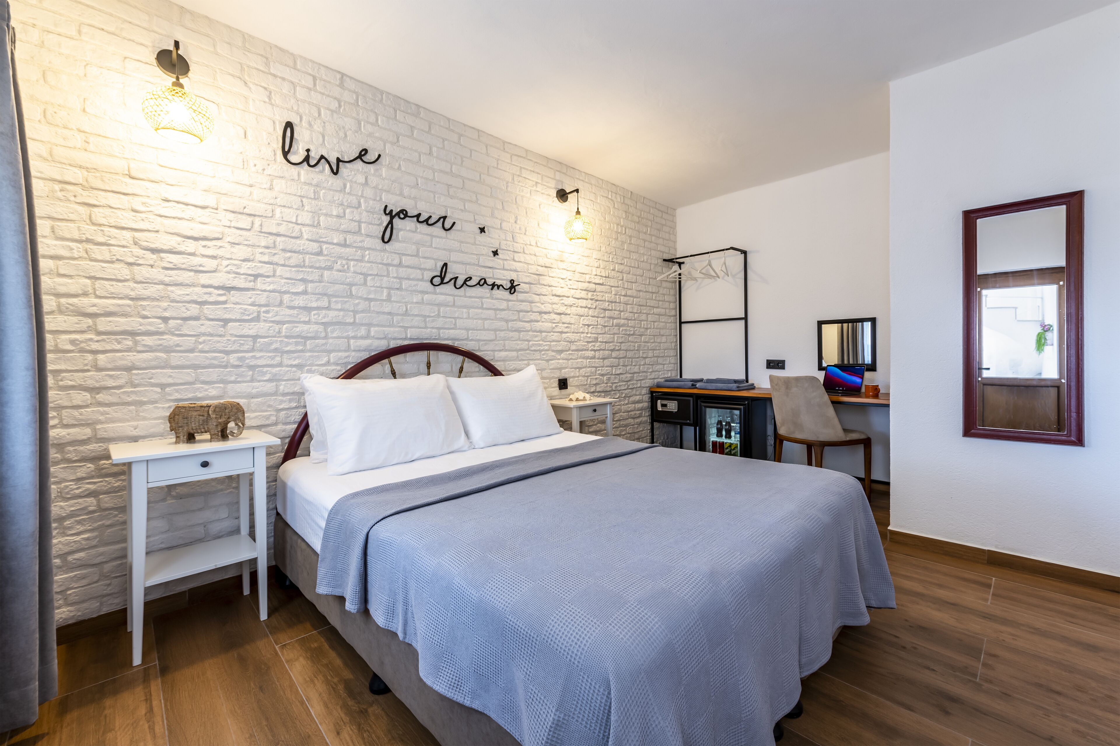 Een slaapkamer met een witte bakstenen muur, een bed met wit beddengoed, een nachtkastje, een bureau met een stoel, en een spiegel aan de muur.