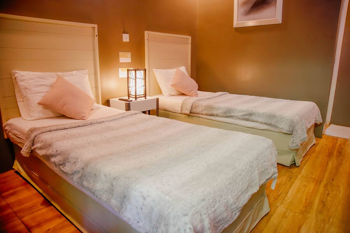 Una habitación con dos camas, cada una con cabecero y manta blancos. Entre las camas, una mesita de noche con una lámpara.