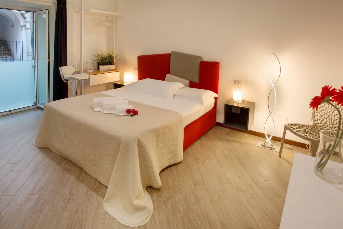 Una habitació d'hotel amb llit, taules de nit, una cadira, una làmpada i vista a un edifici a l'exterior.