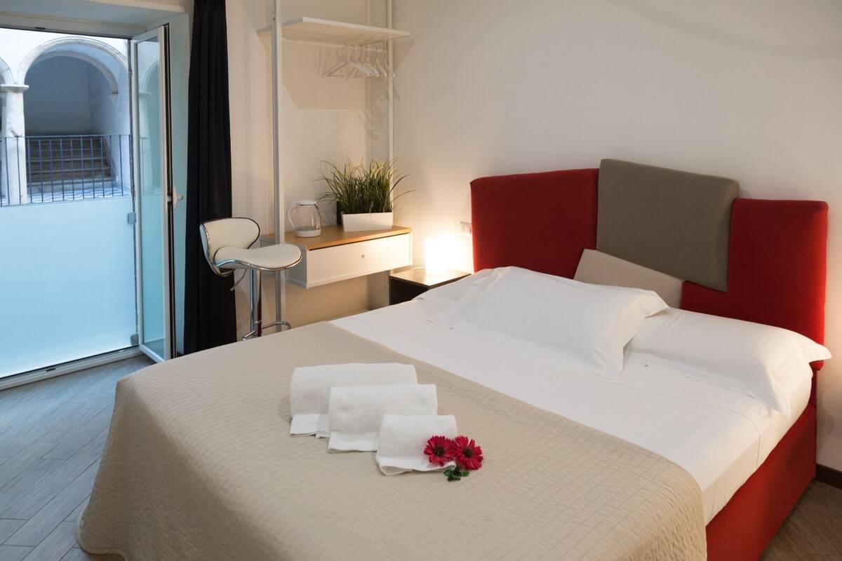 Habitació d'hotel amb llit, escriptori amb cadira, planta en test i vista a un balcó.