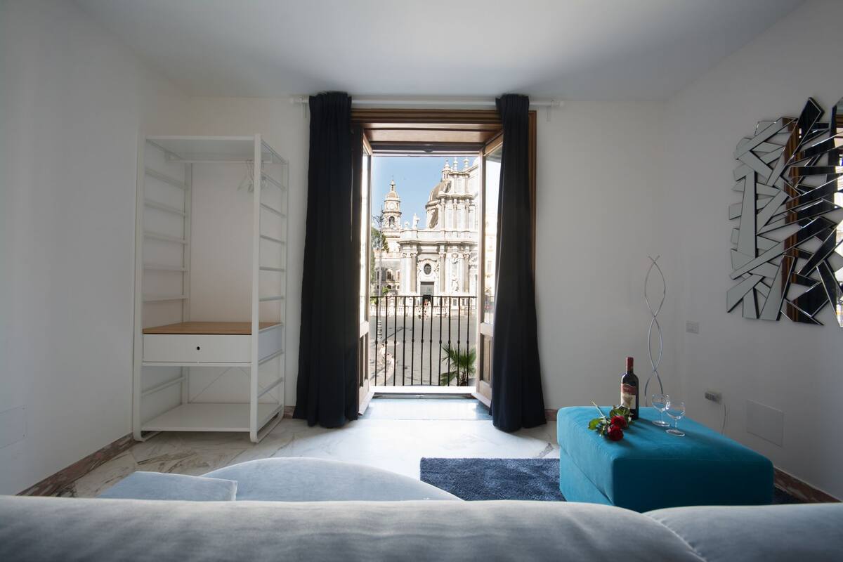 Habitació amb balcó, armari blanc, otomà blau i mirall decoratiu.