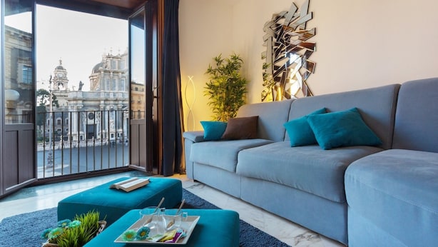 A modern living room with a grey sofa, teal cushions, and a balcony with a view of historic buildings. - Le suites del duomo house (Catania)