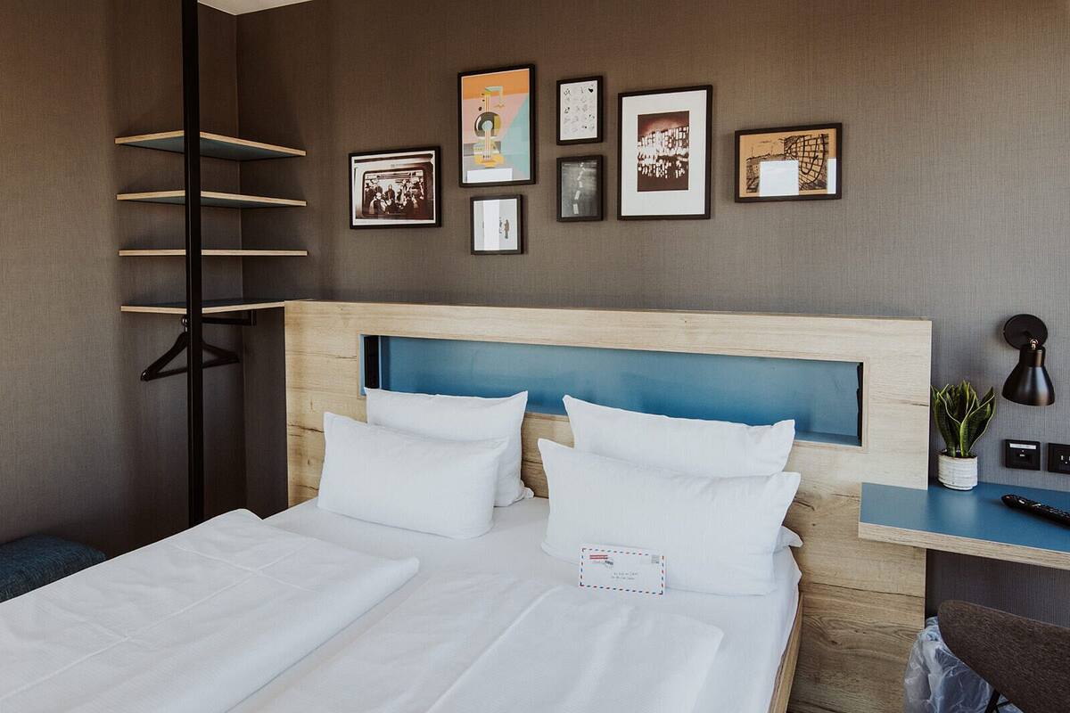 A hotel room with a bed, a desk with a lamp, a plant, and framed pictures on the wall.