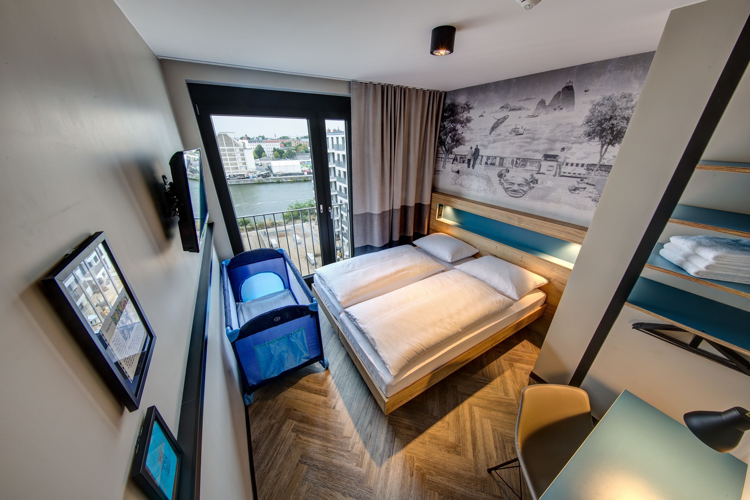 Ein Hotelzimmer mit Bett, Schreibtisch, Stuhl, Koffer und Blick auf eine Stadtlandschaft durch ein großes Fenster.