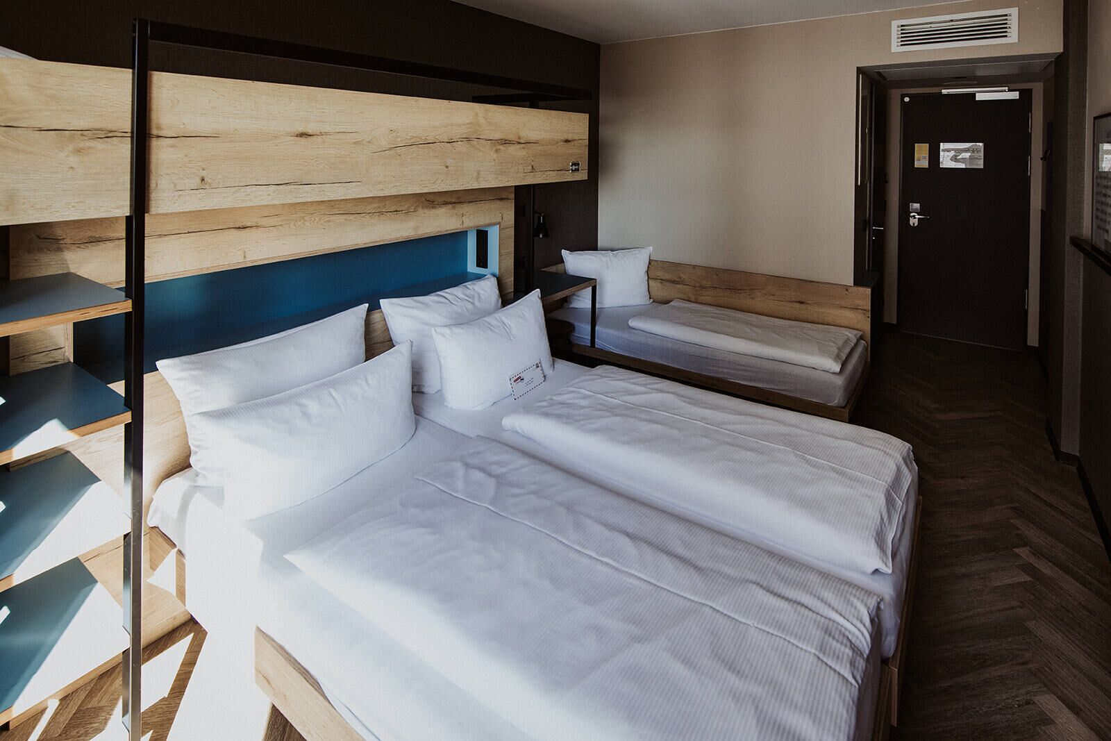 Ett hotellrum med en våningssäng och en enkelsäng, båda med vita sängkläder. Det finns en träpanel vid sänggaveln, ett litet bord och en bänk.