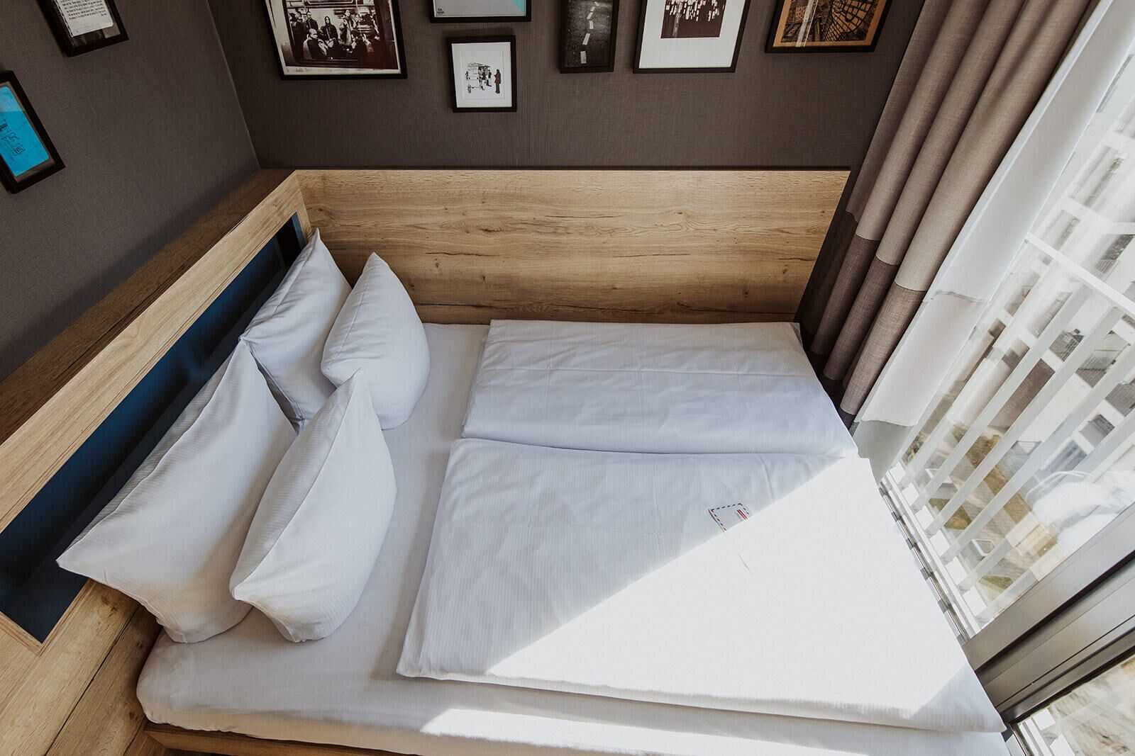A neatly made bed with white bedding in a room with a wooden headboard, framed pictures on the wall, and a window with curtains.