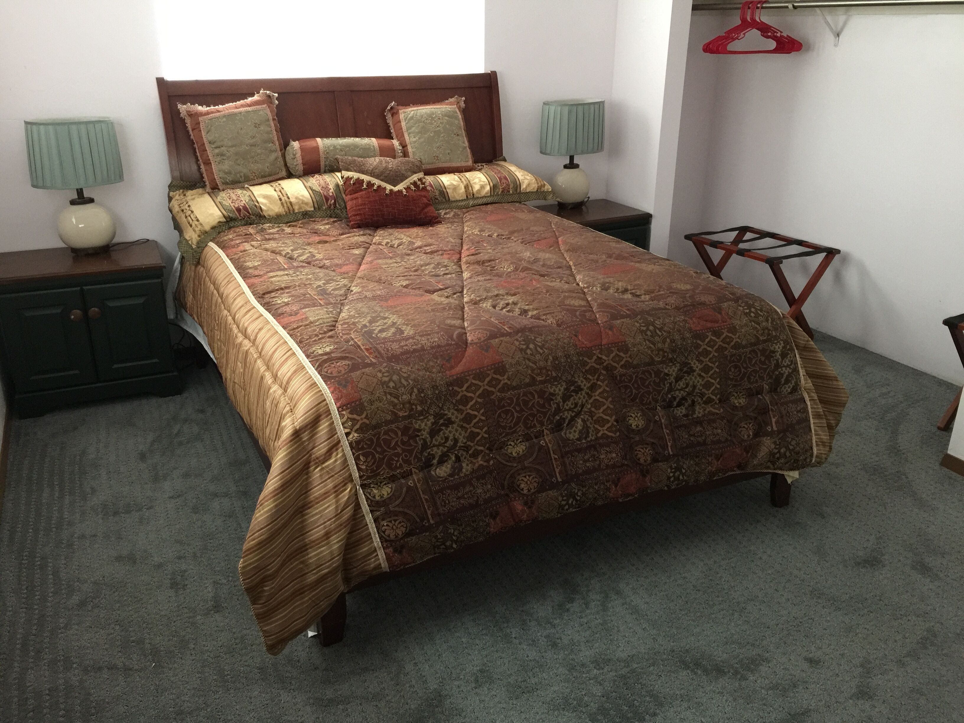 Ein ordentlich bezogenes Bett mit gemustertem Überwurf und Kissen, zwei Nachttische mit Lampen, ein Holzstuhl und ein roter Kleiderhaken an der Wand.